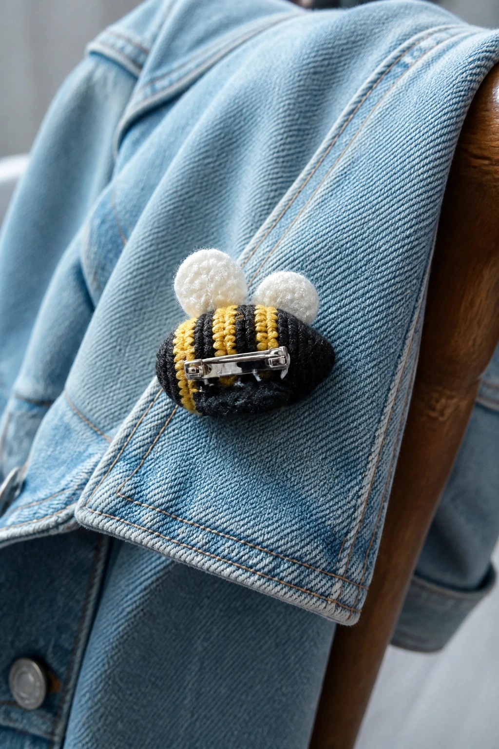 Crocheted bee with black and yellow stripes and white pom-pom wings pinned to blue denim fabric.