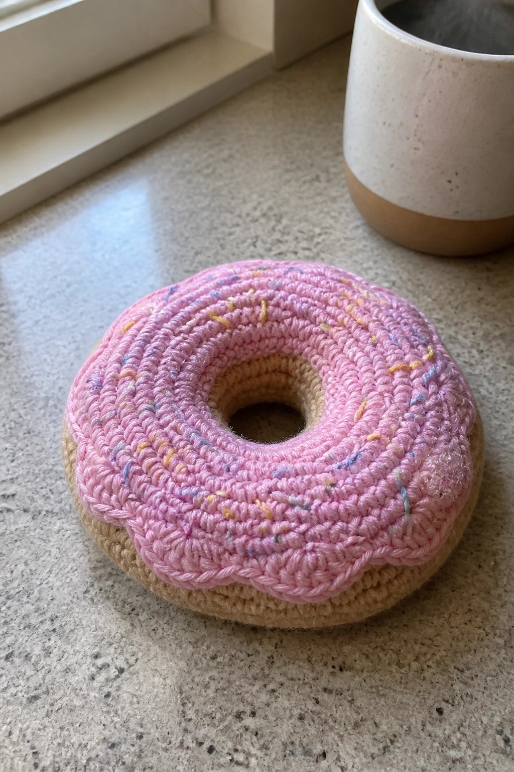 Pink crocheted donut with colorful sprinkles on a speckled kitchen countertop.
