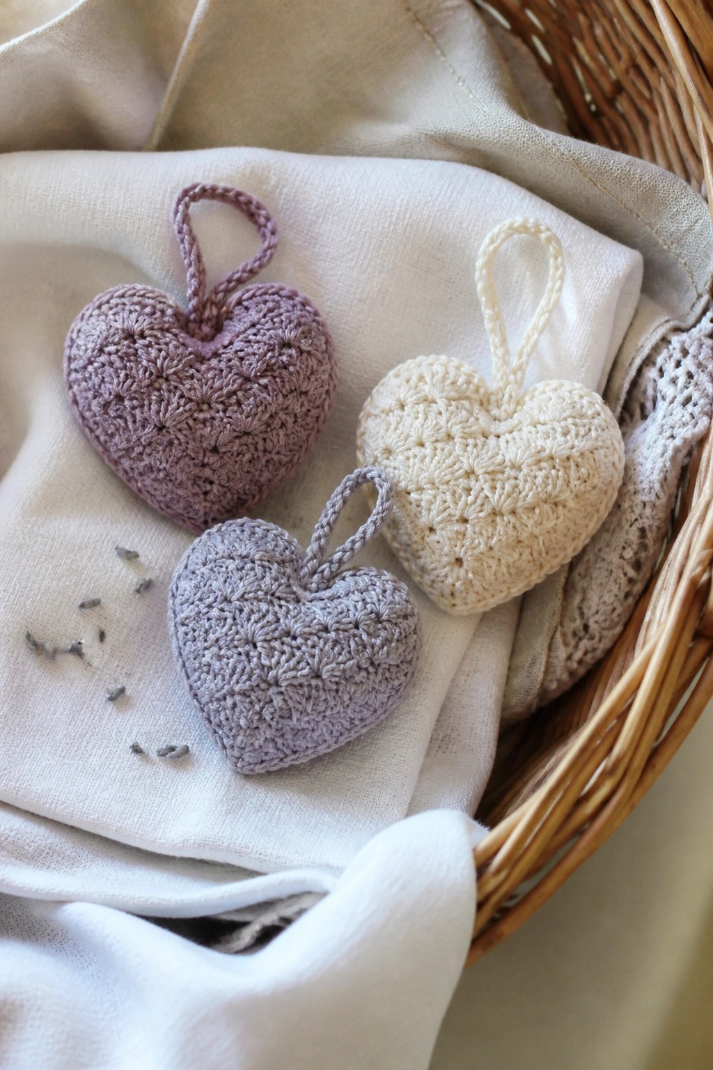 Three crocheted hearts in light purple, cream, and gray with hanging loops on white fabric inside a basket.