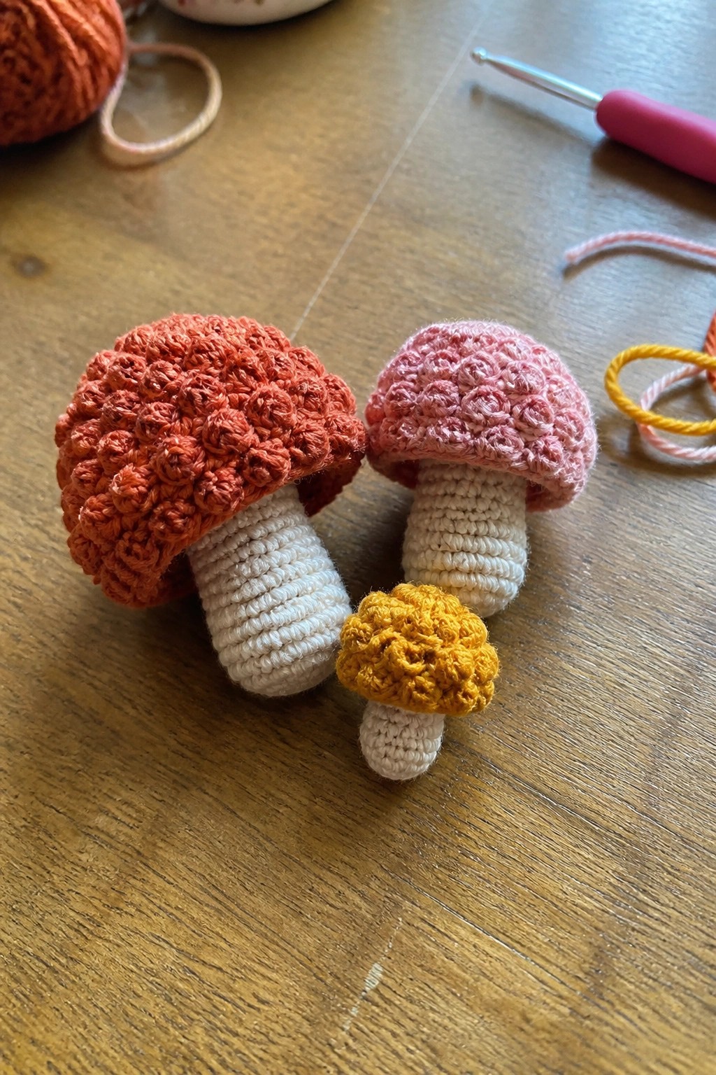 Three crocheted mushrooms in orange, pink, and yellow with white stems on wood.