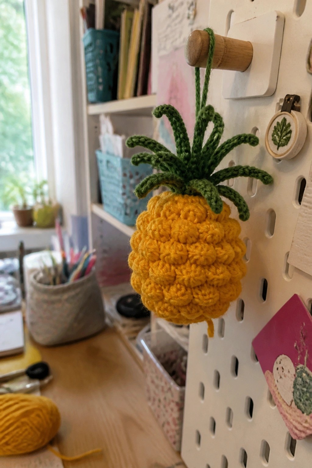 Yellow crocheted pineapple ornament with green top hanging from pegboard peg.