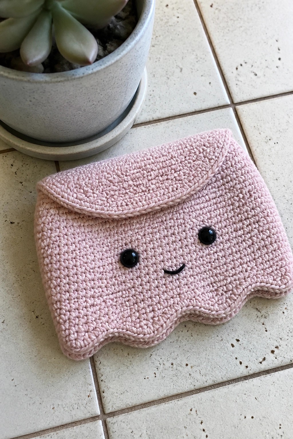 Pink crocheted pouch with smiling face beside potted succulent on tiled floor