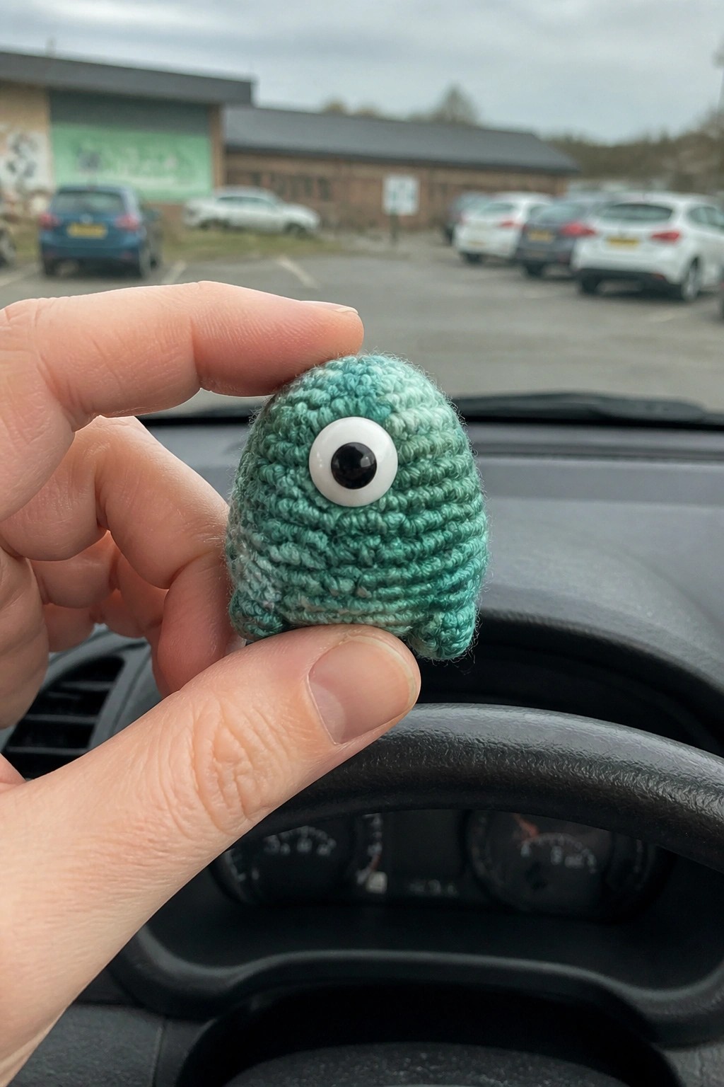Hand holding small teal crocheted one-eyed plush toy inside car with parking lot view.