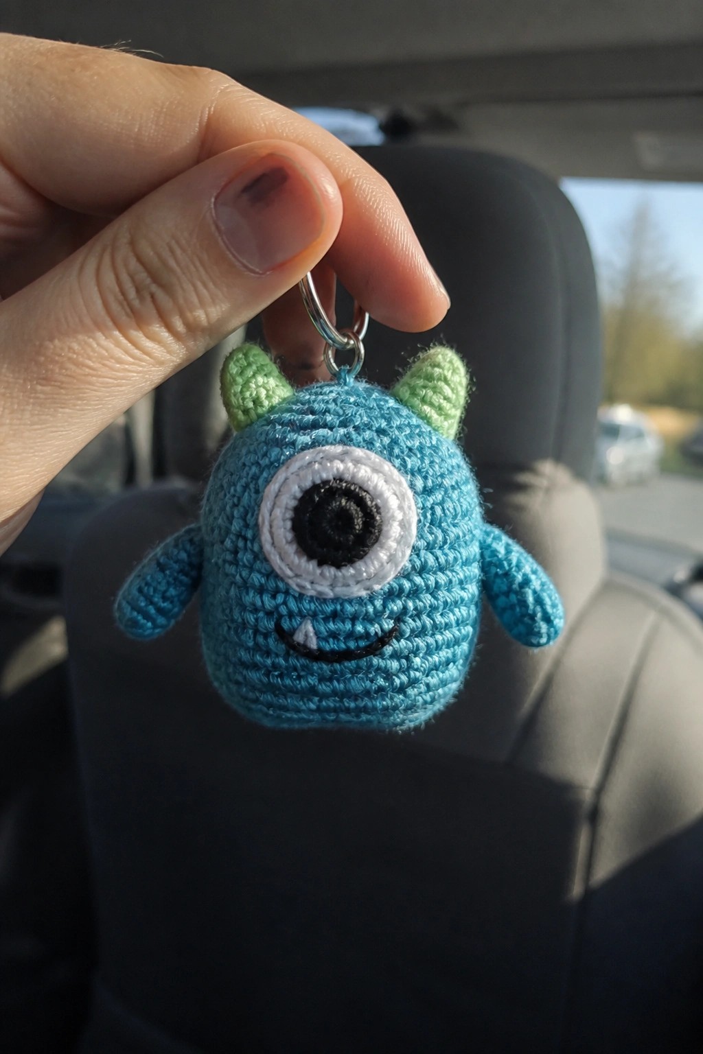 Hand holding blue crocheted one-eyed monster keychain with green horns in car