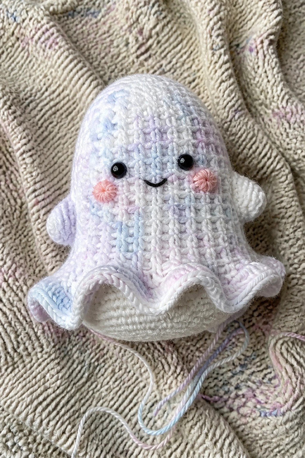 Crocheted pastel ghost amigurumi with smiling face on knitted blanket