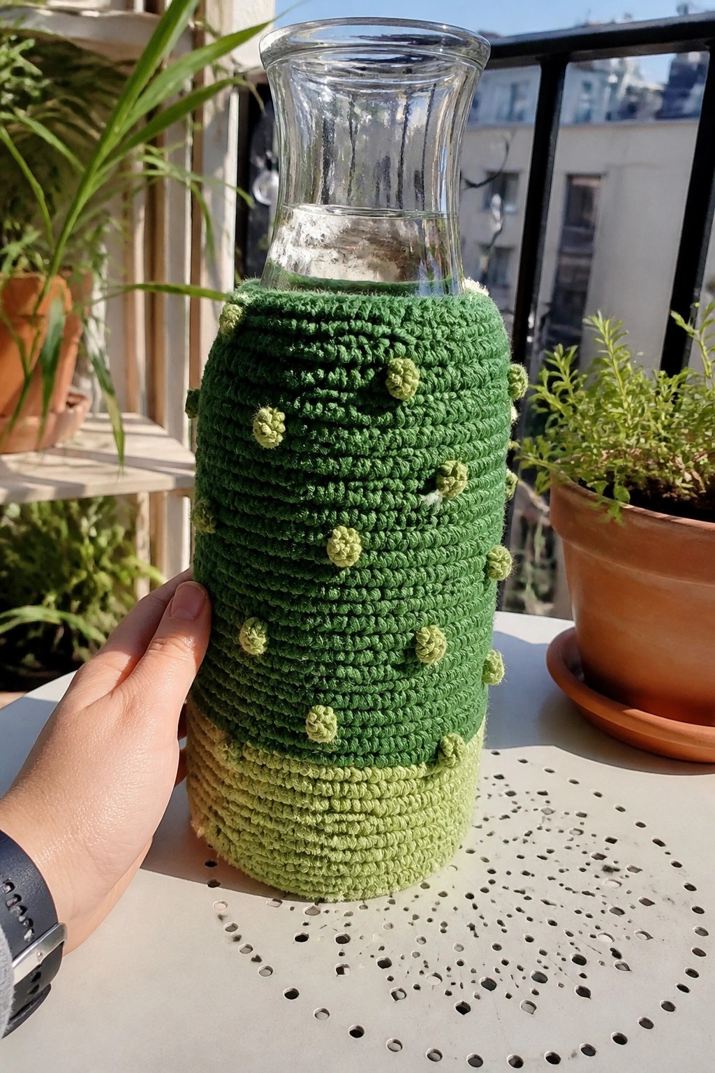 Hand holding green crocheted cactus cover on clear glass vase with pom-poms outdoors
