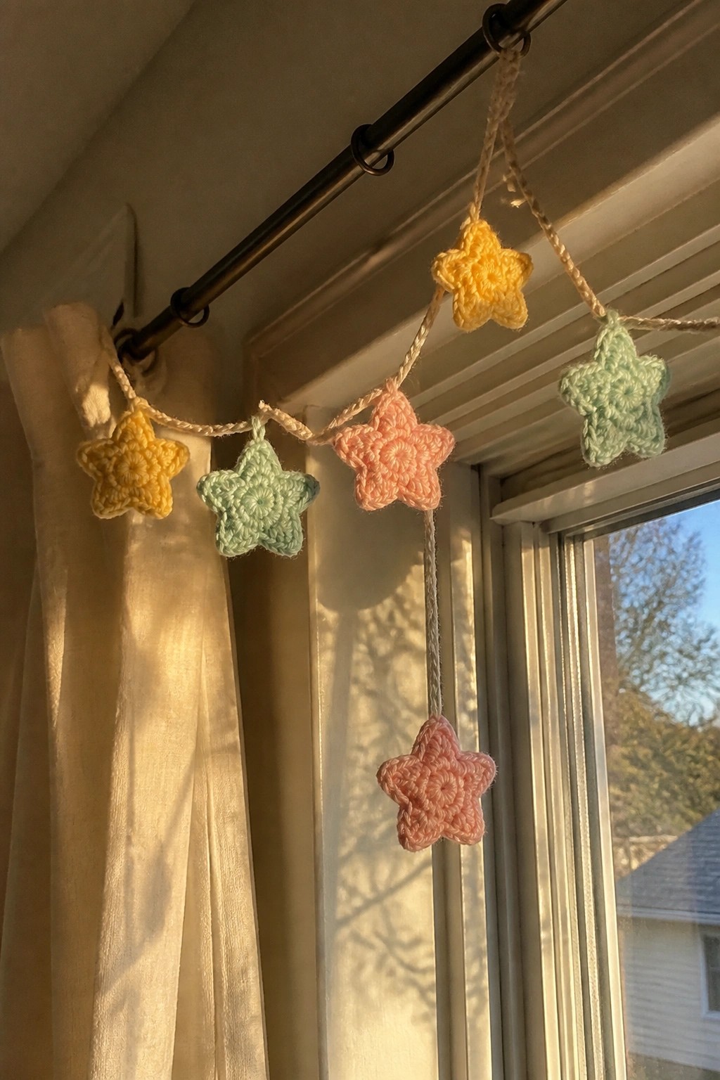 A string of small crocheted stars in yellow, mint, and peach hangs from a black curtain rod beside a window.