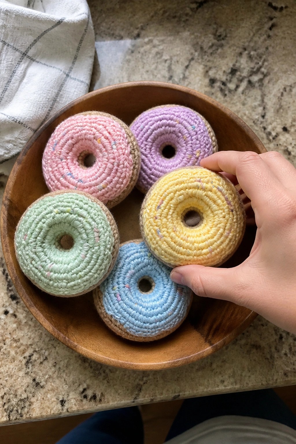 Hand lifting yellow crocheted donut from bowl of pastel ones on marble.