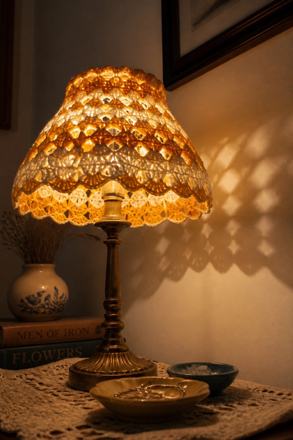 Glowing crocheted lampshade on brass lamp with books, bowls, and jewelry nearby.