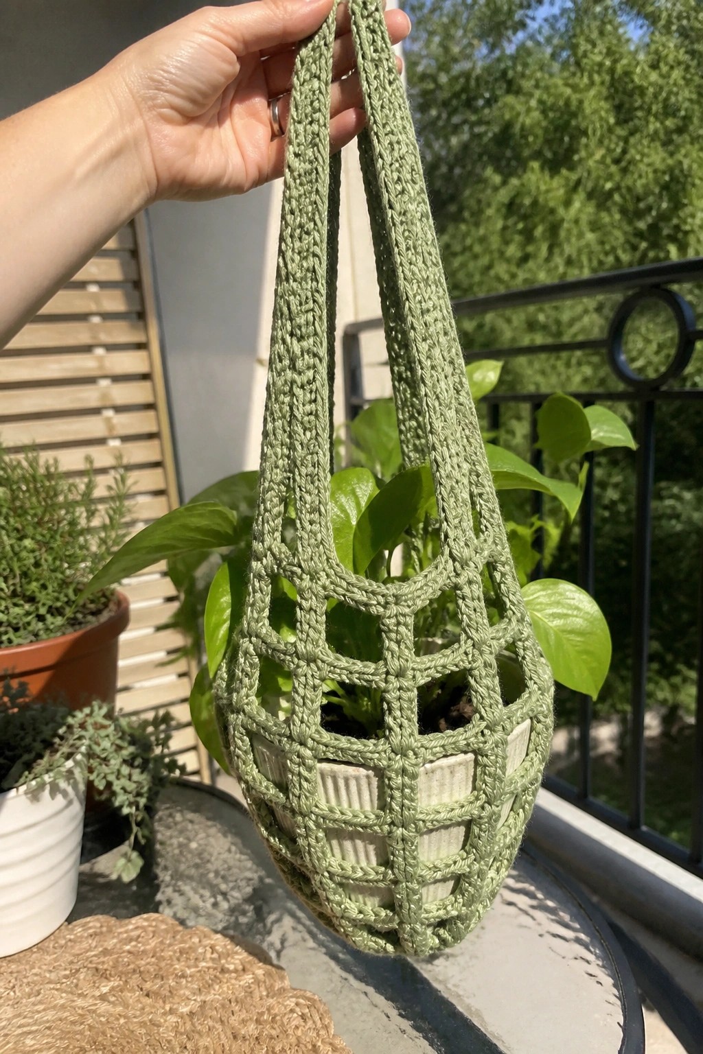 Hand holding sage green crocheted hanging planter with leafy potted plant outdoors.