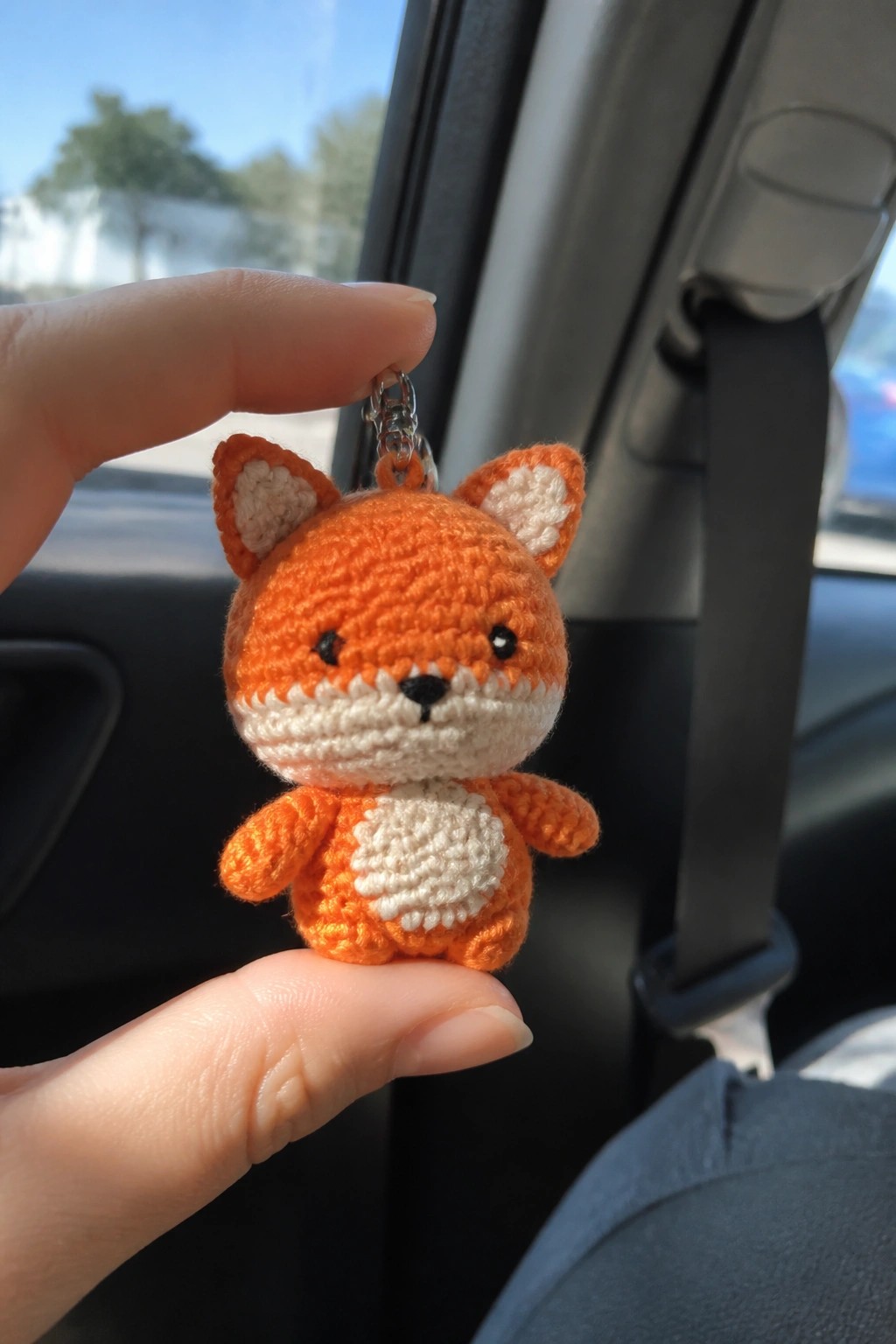 Hand holding small orange crocheted fox keychain with white belly inside car.