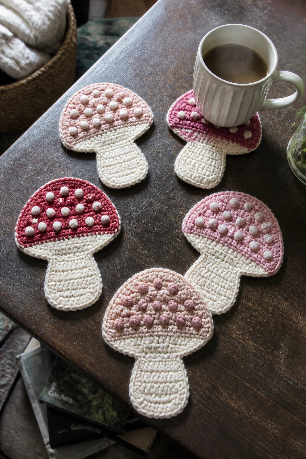 Pink and white crocheted mushroom coasters on dark wooden table with white mug.