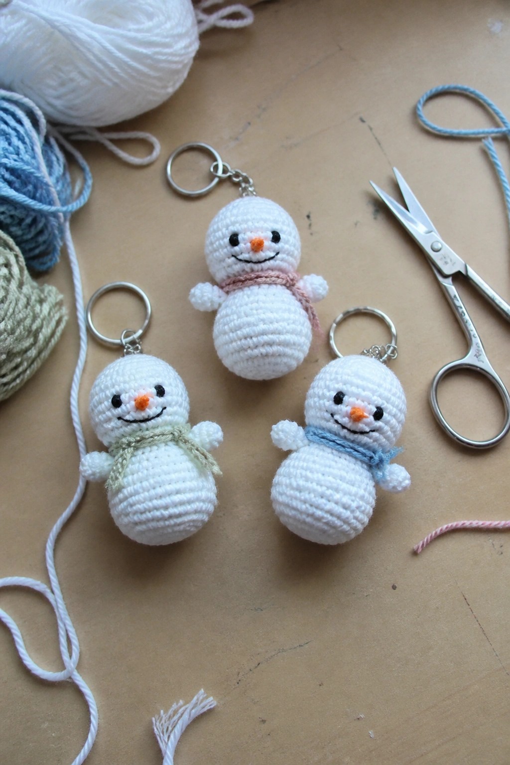 Three crocheted snowman keychains with pink, green, and blue scarves amid yarn and scissors.