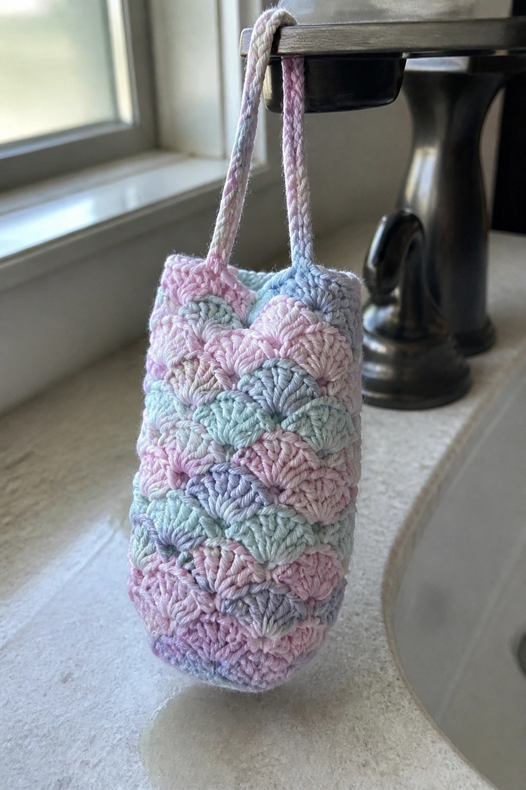 Pastel crocheted pouch with braided strap hanging from bathroom sink faucet