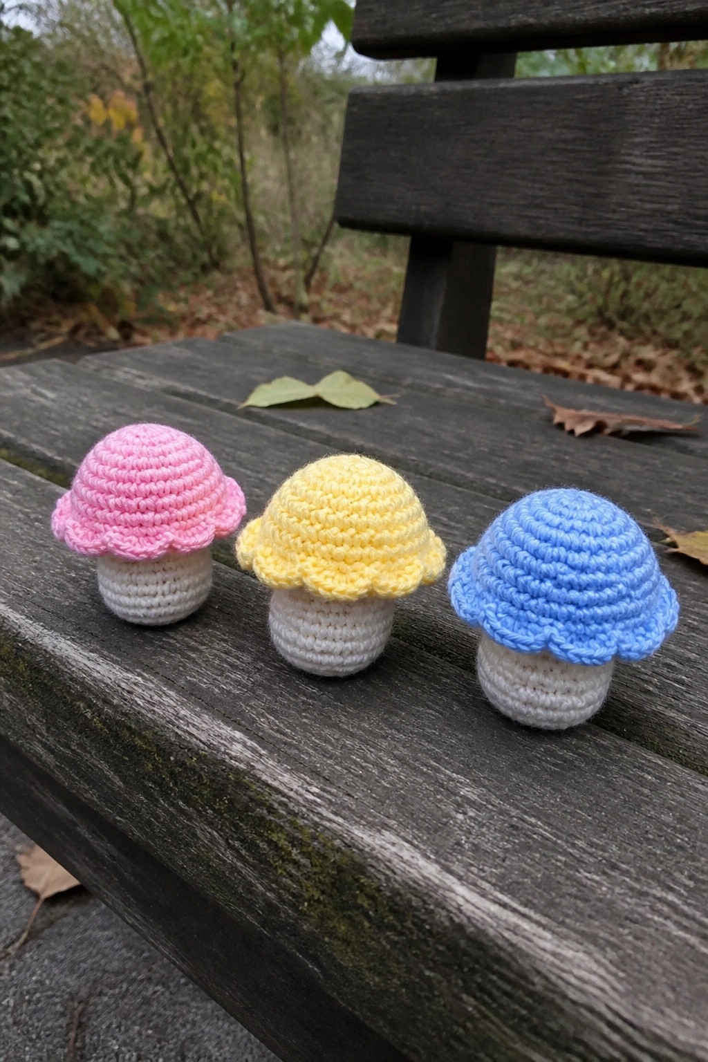 Three colorful crocheted mushrooms with pink, yellow, and blue caps on a bench.