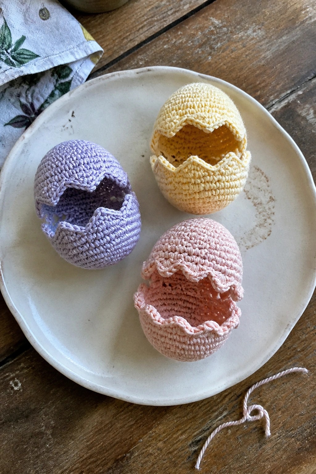 Three pastel crocheted eggs with cracked tops on a white plate on wood.