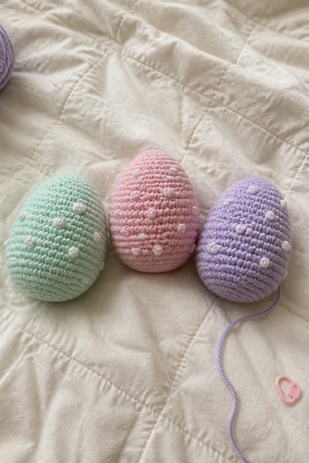 Three pastel crocheted eggs in mint, pink, and lavender with white dots on quilted fabric.