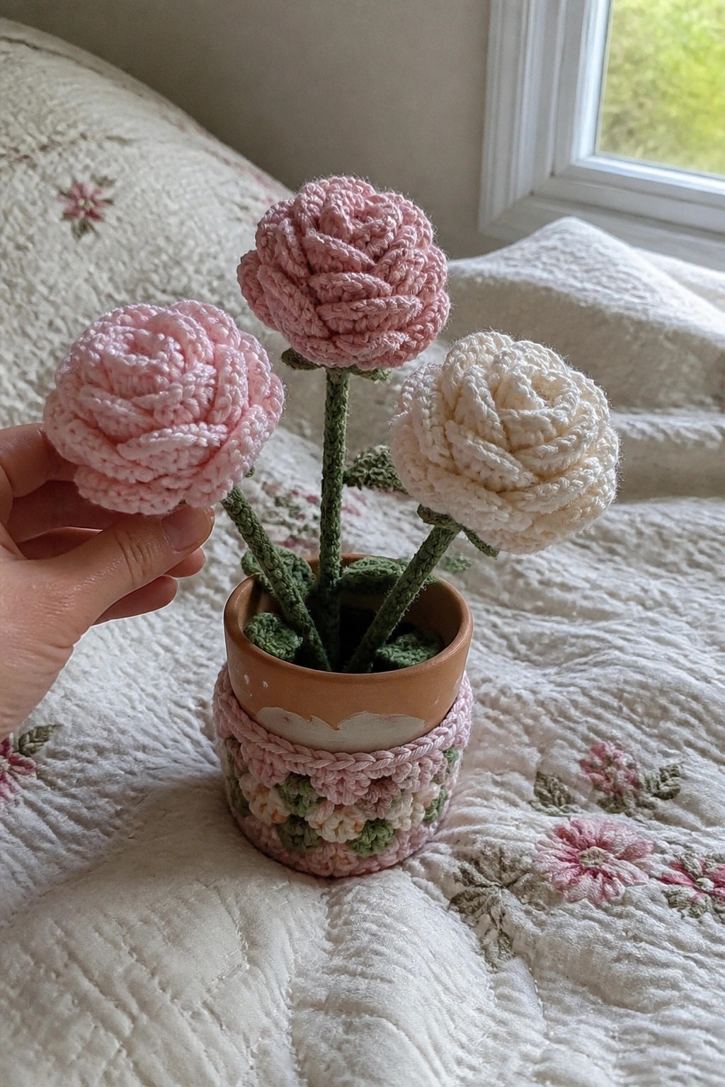 Hand holding pink crocheted rose beside potted pink and white flowers on quilt