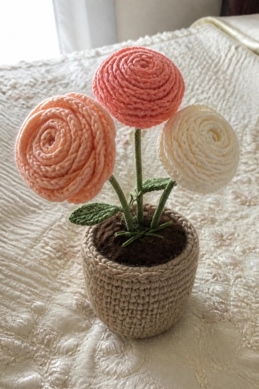 Three crocheted spiral flowers in peach and white inside a beige yarn pot with green stems.