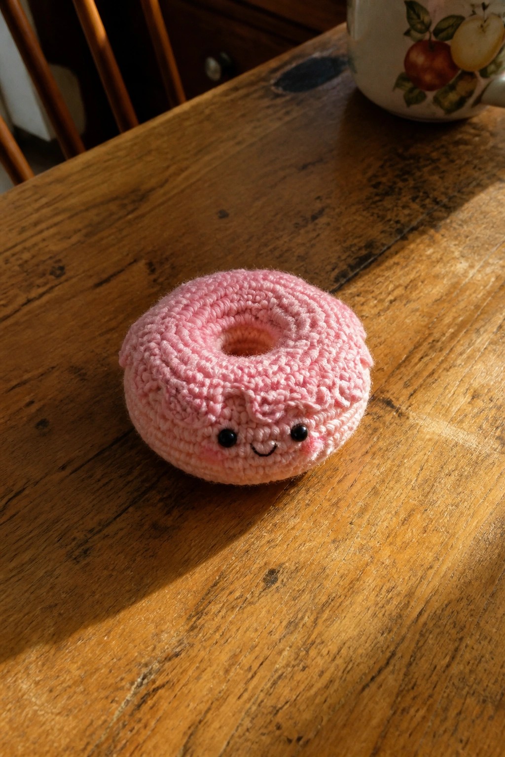 Pink crocheted donut with smiling face on sunlit wooden table.