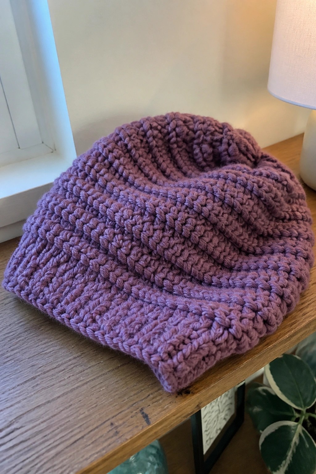 Thick purple chunky knit cowl coiled on wooden table near lamp