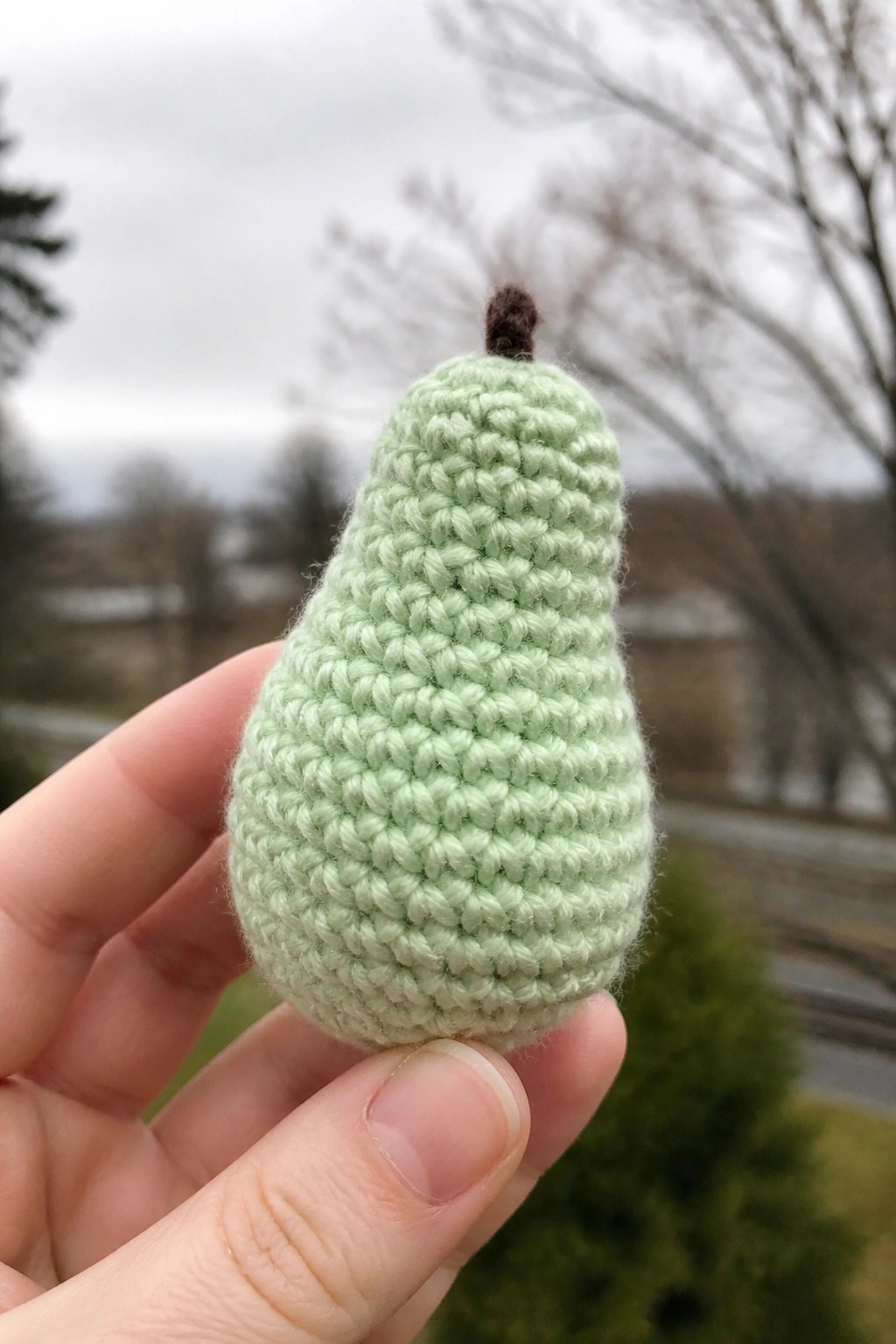 Hand holding light green crocheted pear with brown stem against blurred background.