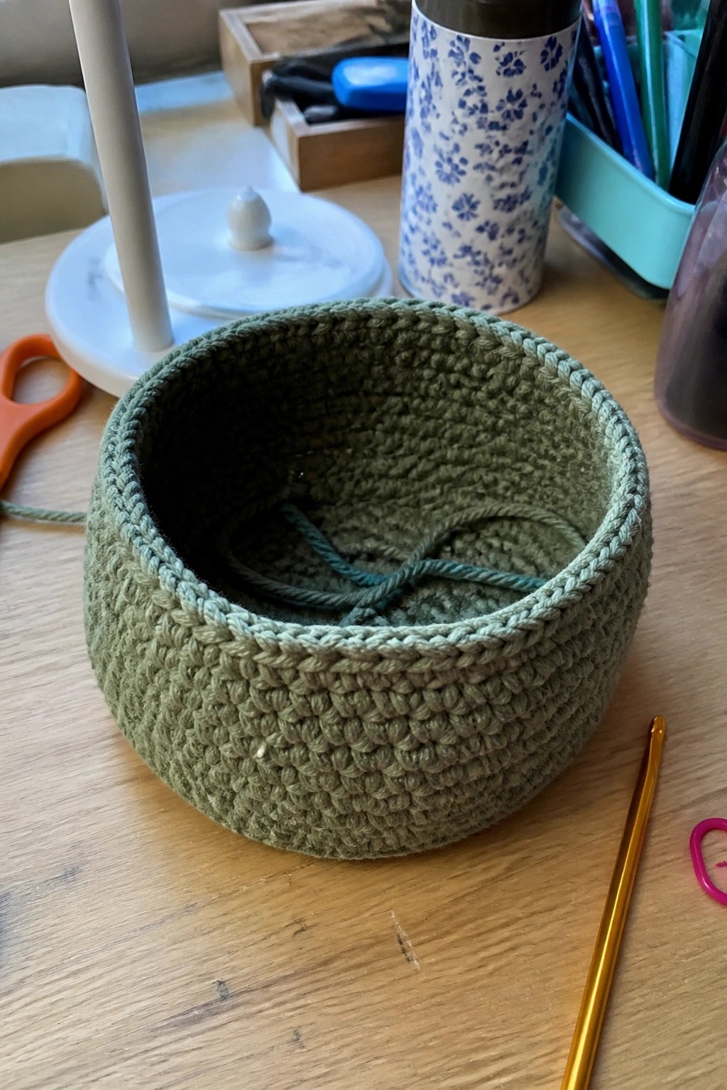 Green crocheted basket with loose yarn on wooden table beside gold hook