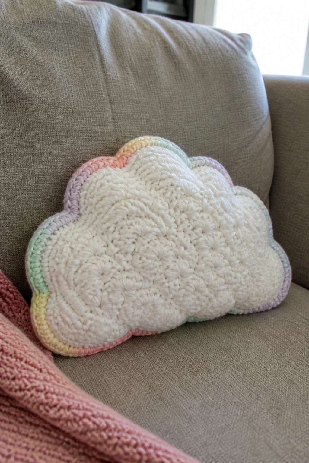 Crocheted white cloud pillow with pastel rainbow border on gray couch