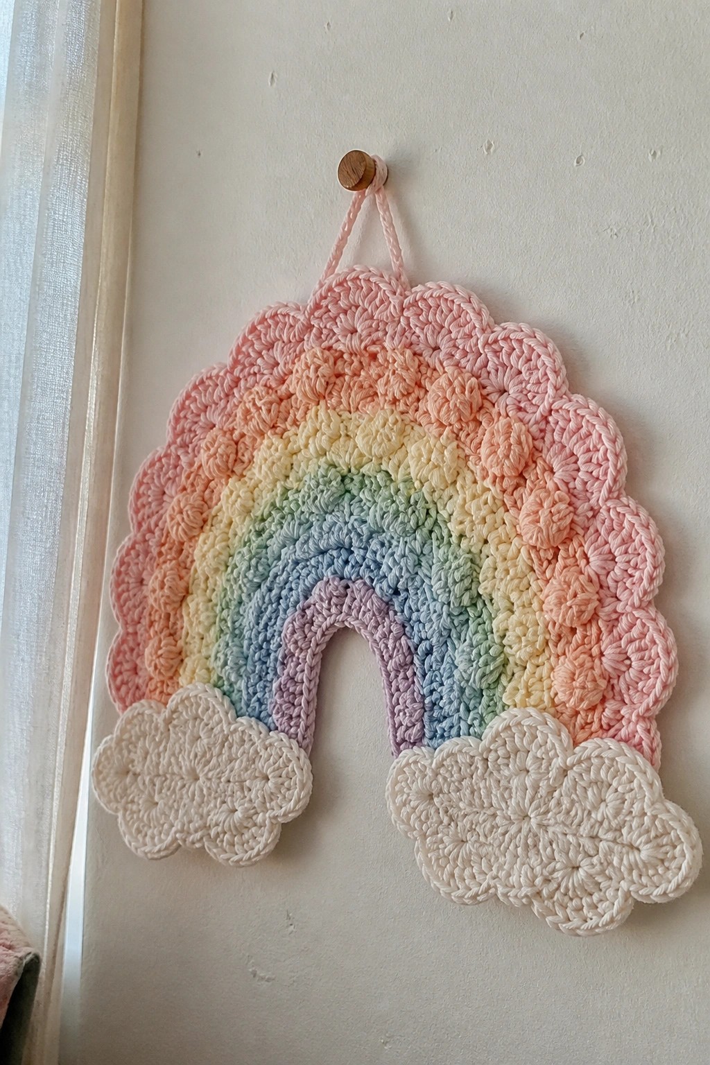 Crocheted pastel rainbow with white clouds hanging from wooden peg on wall.