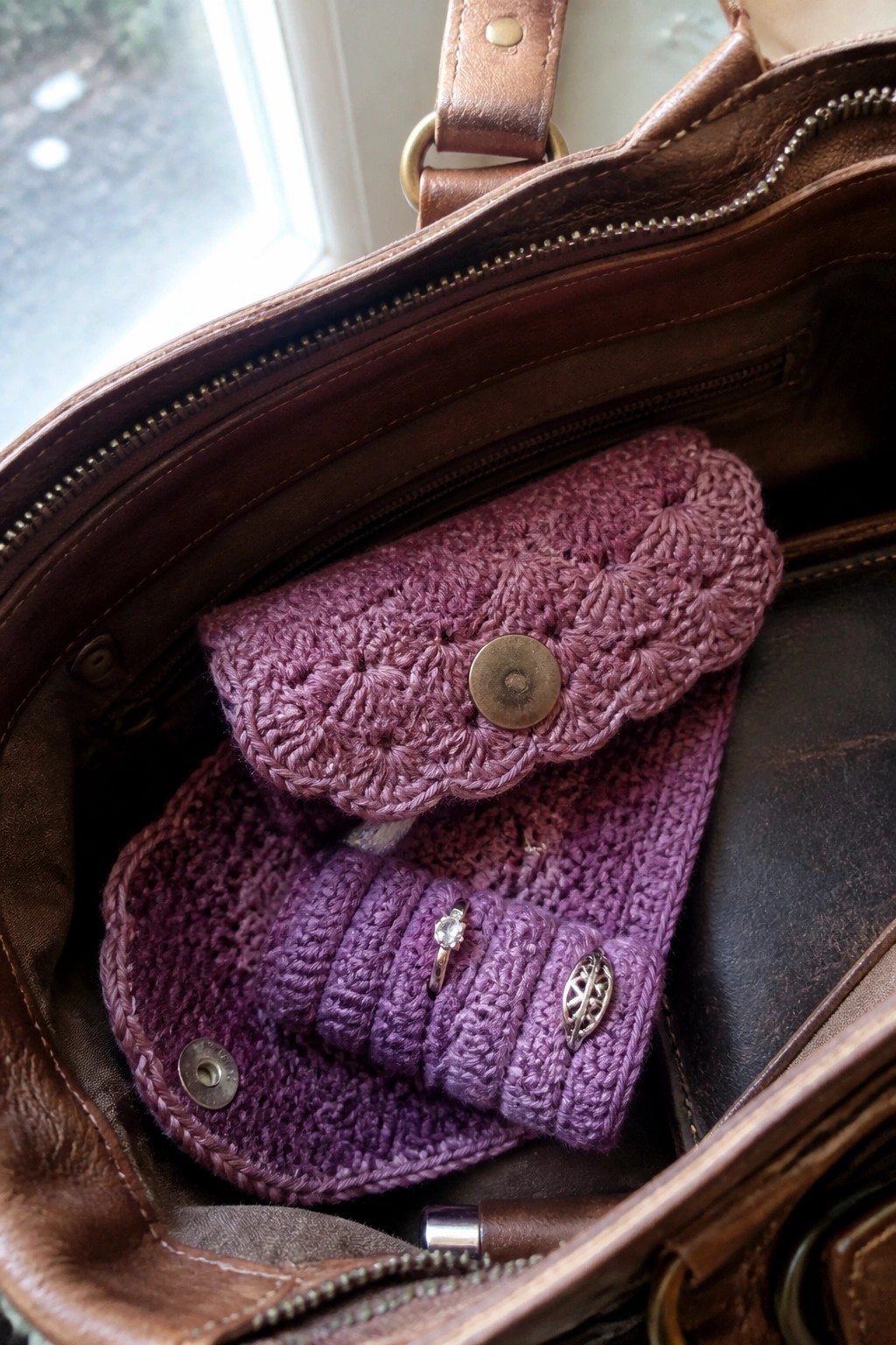 A purple crocheted pouch with a button sits inside an open brown leather bag next to another purple crocheted item holding a ring.