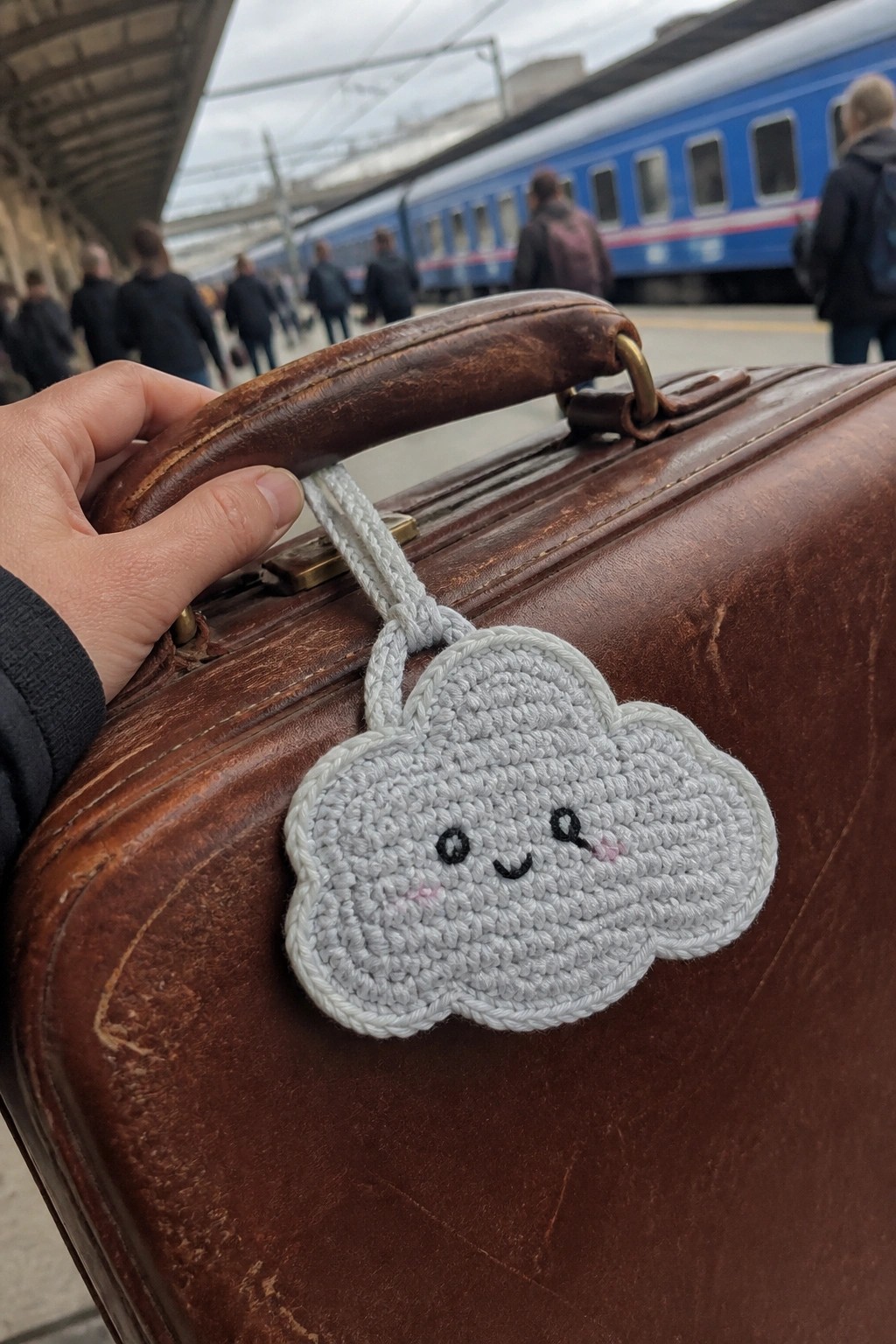Hand holding brown leather suitcase with smiling crocheted cloud charm at train station