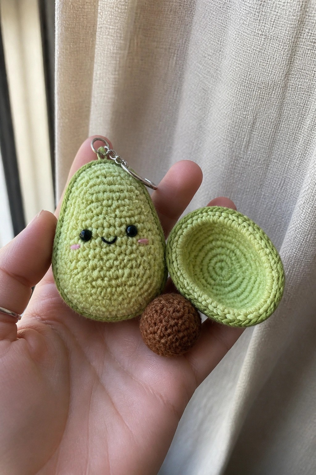 Hand holding light green crocheted avocado keychain with face, pit, and half.