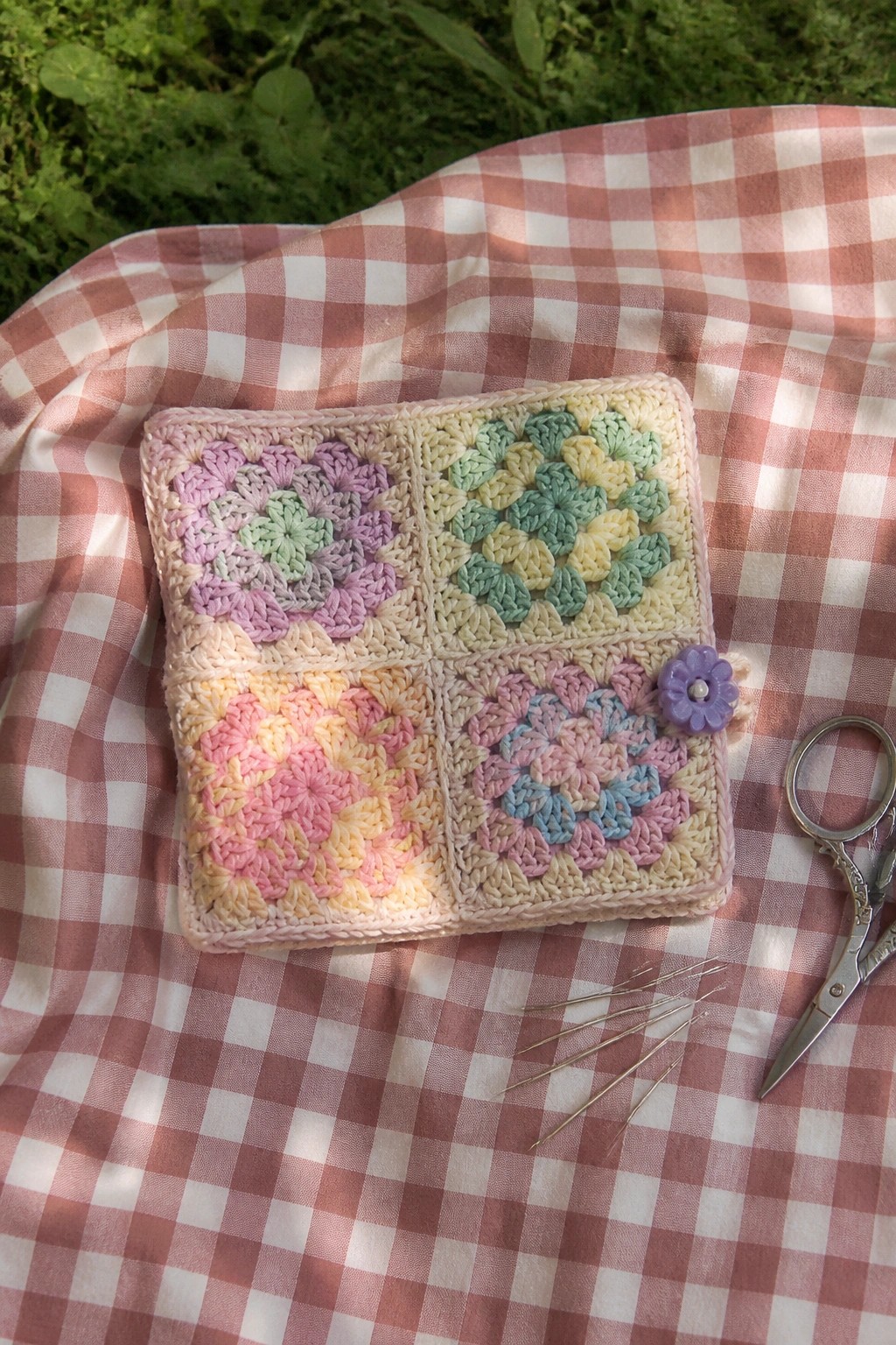 Pastel crocheted granny square pouch on pink checkered fabric with scissors and needles.