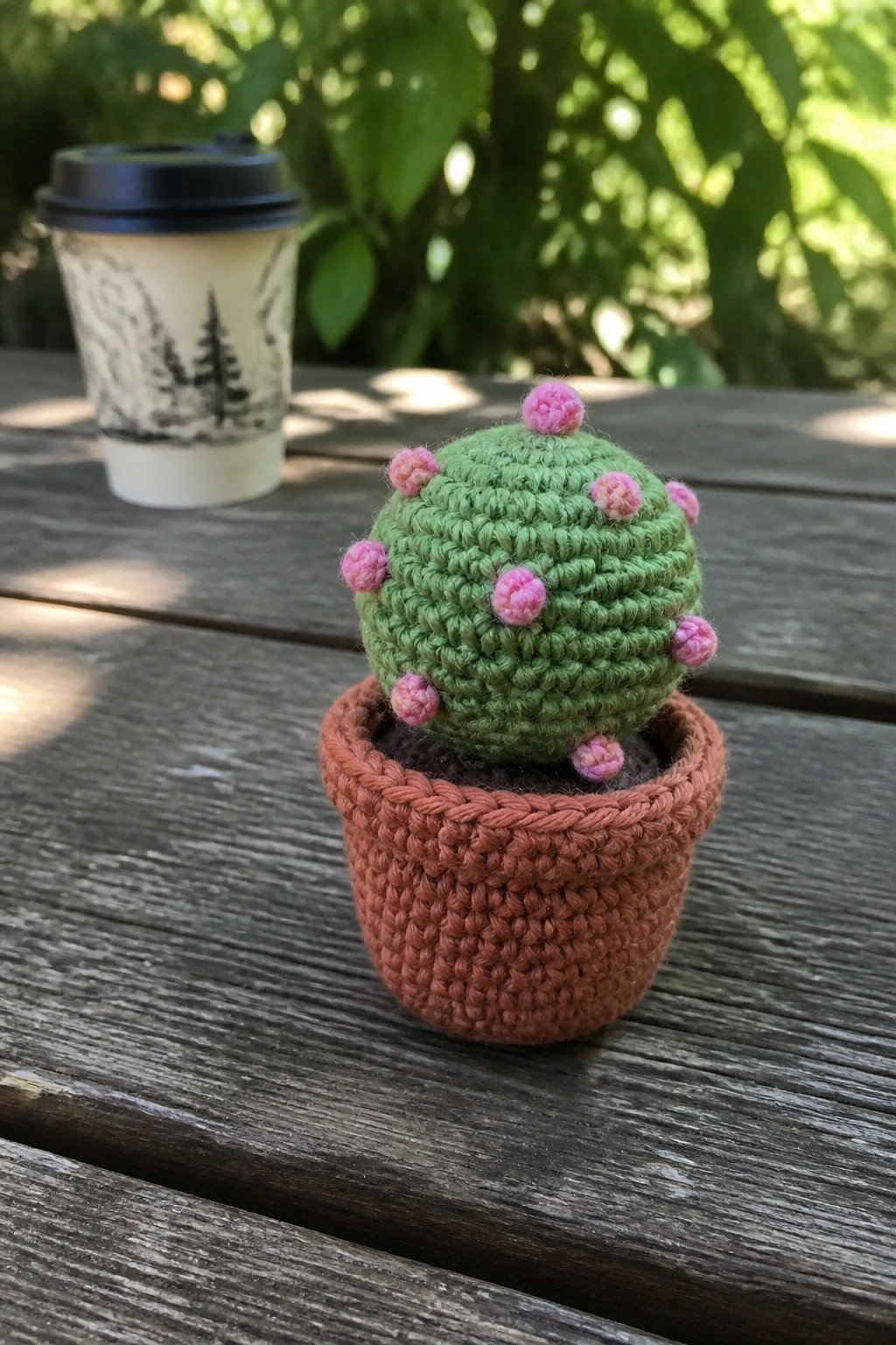 Green crocheted cactus with pink buds in brown pot on wooden table near coffee cup.