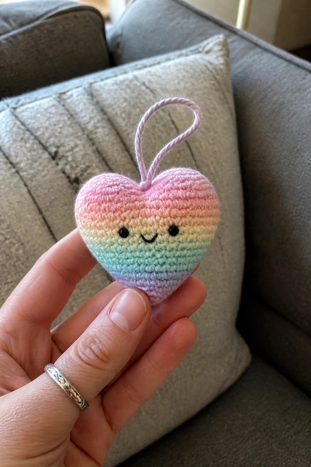 Hand holding pastel rainbow crocheted heart ornament with smiling face and loop.