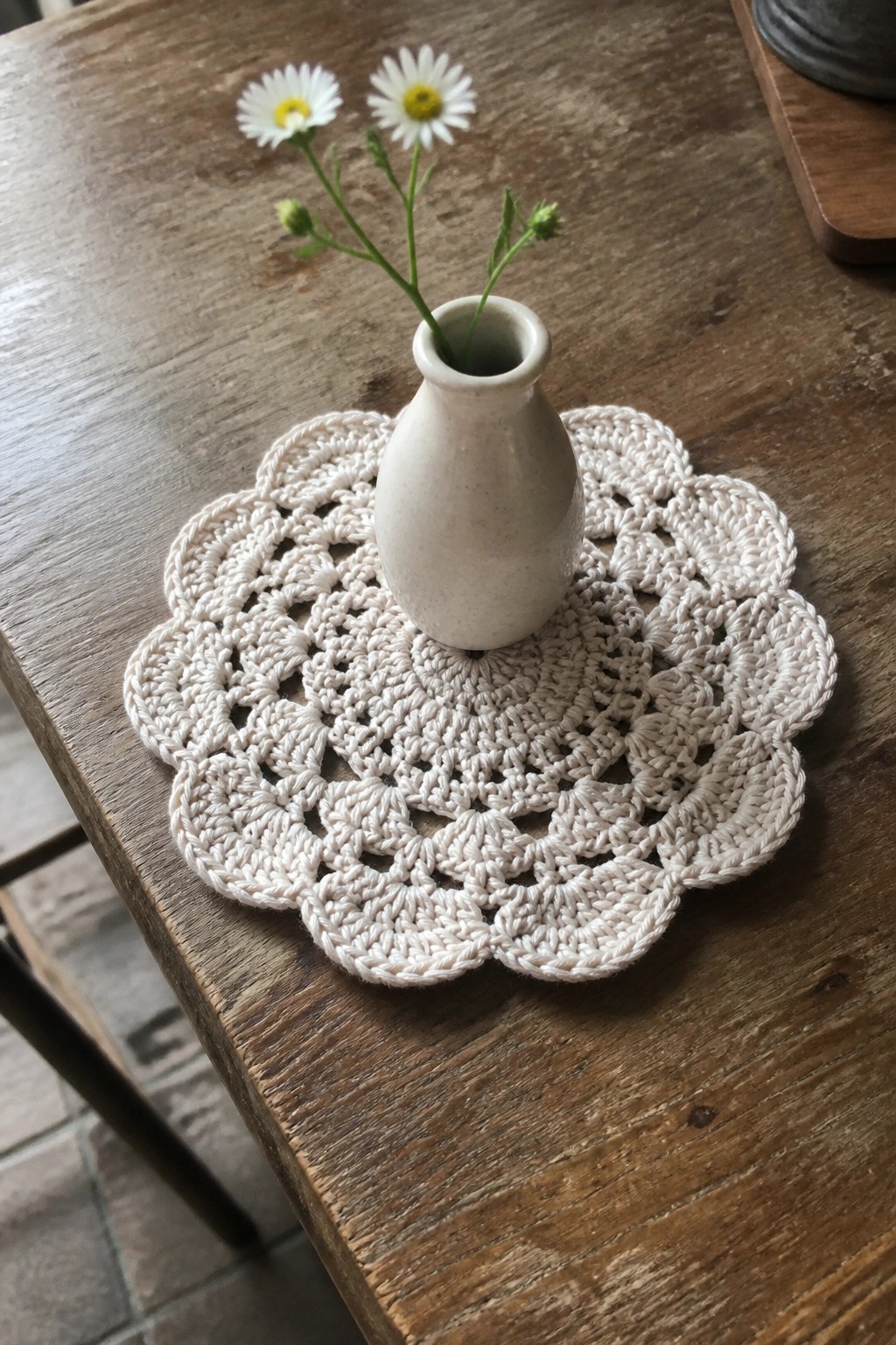 Small white vase with daisies on crocheted doily atop rustic wooden table.