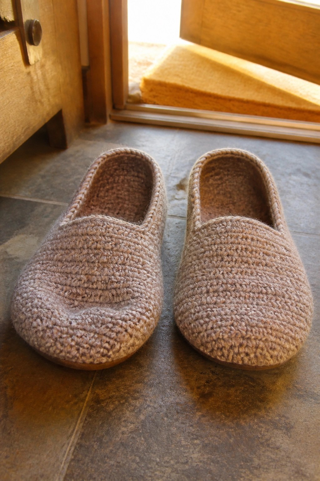 Knitted brown slippers on tiled floor by open wooden door with doormat