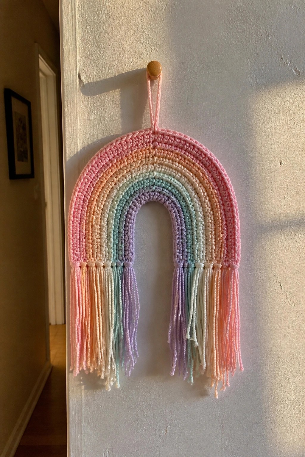 Crocheted pastel rainbow wall hanging with tassels on wooden peg.