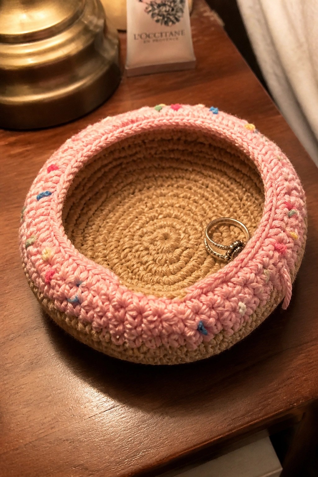 Pink crocheted basket with colorful dots holding a silver ring on wooden table