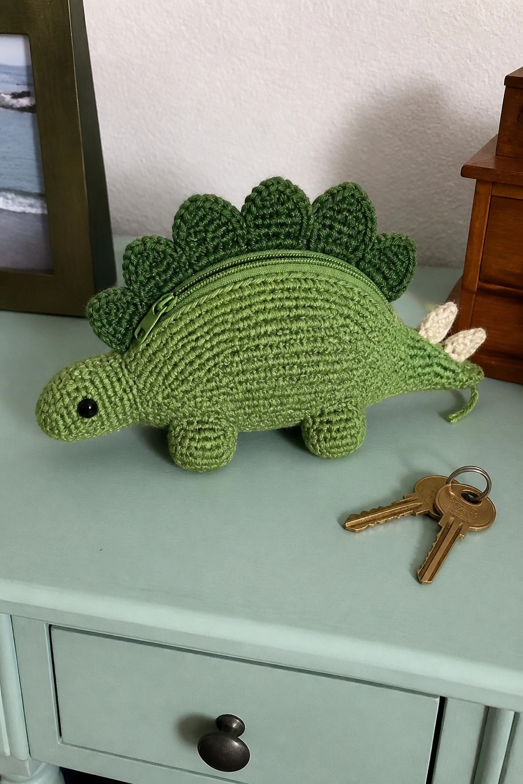 Green crocheted stegosaurus pouch with zipper on light blue table beside keys