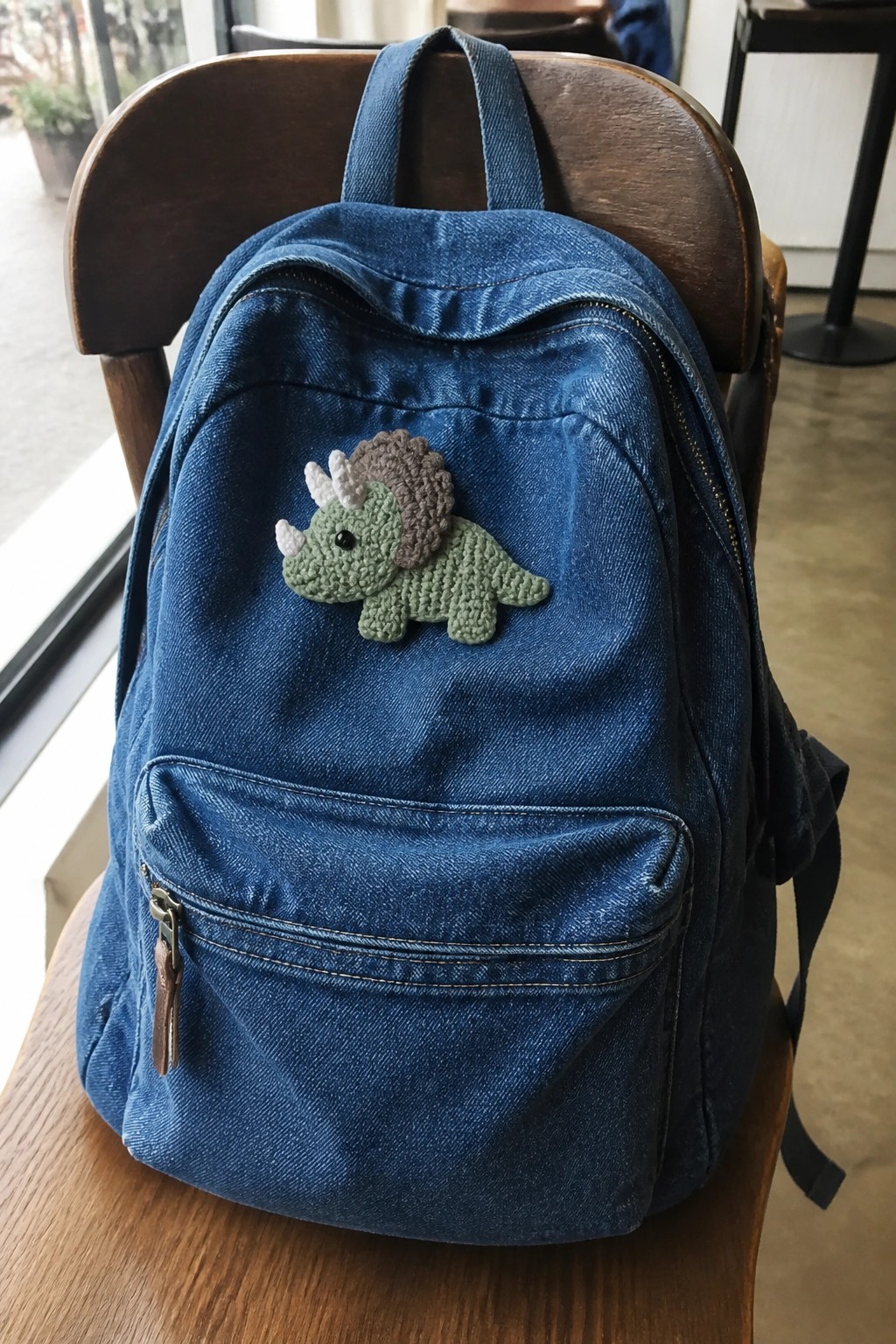 Blue denim backpack with crocheted triceratops patch on wooden chair seat