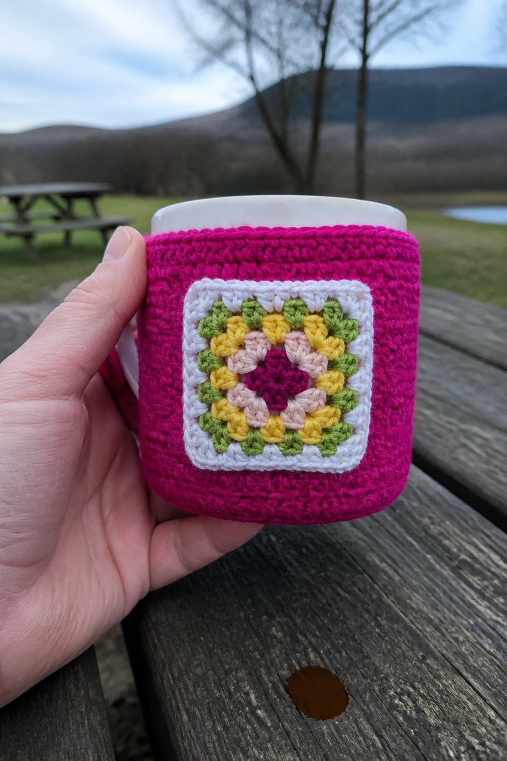 Hand holds white mug in pink crocheted cozy with colorful floral granny square.