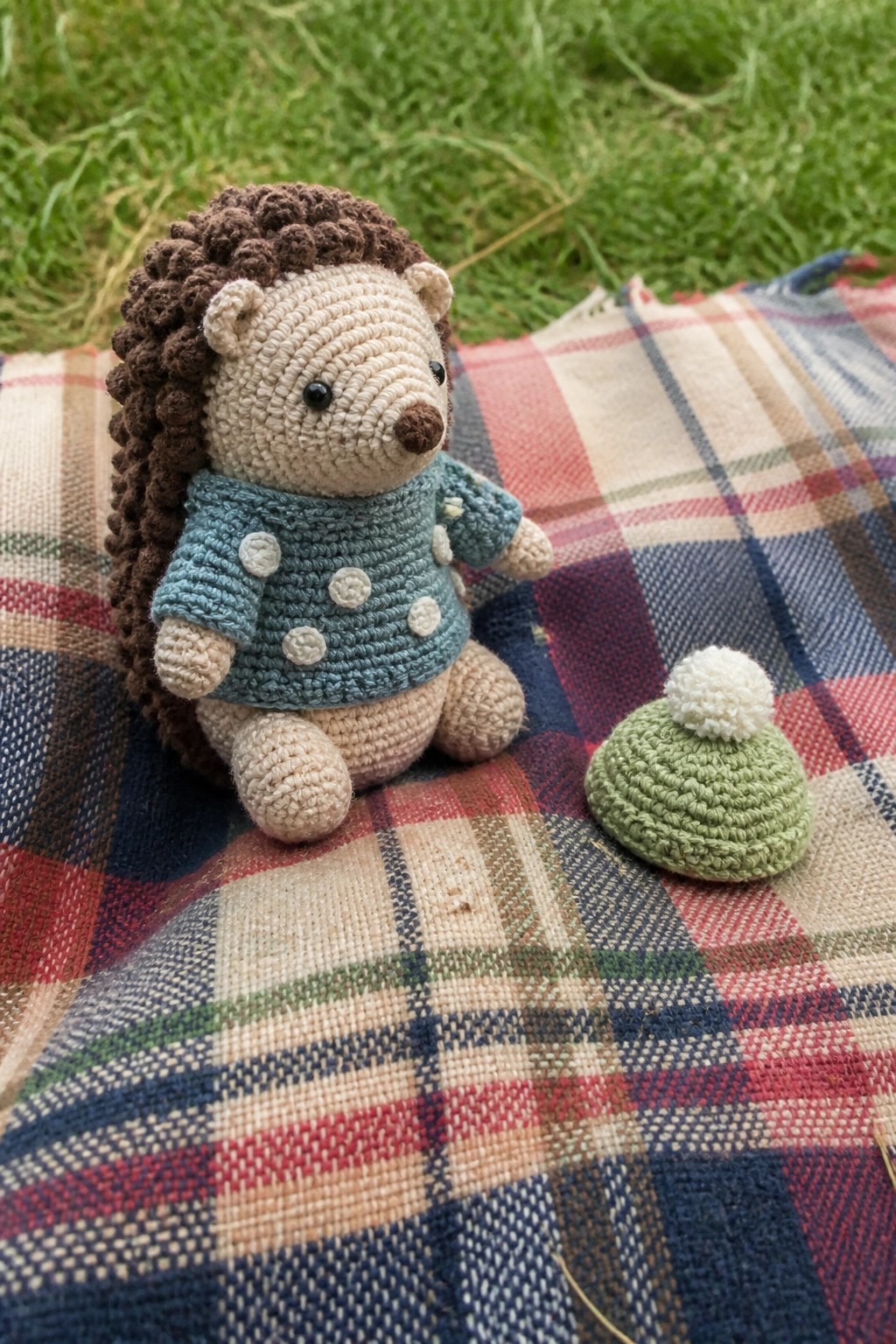 Crocheted hedgehog in blue polka-dot sweater beside green pom-pom hat on plaid blanket