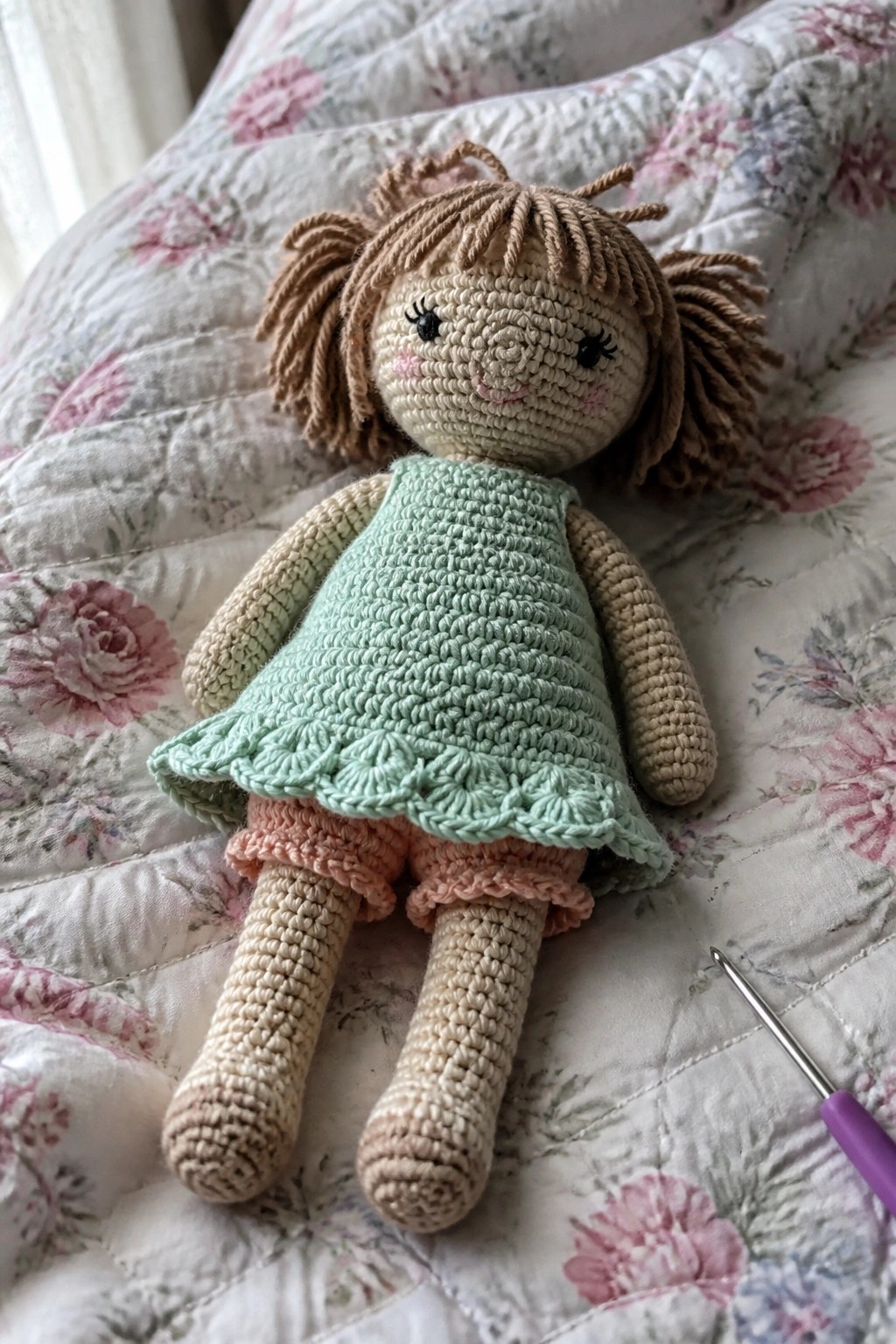Crocheted doll with brown pigtails in mint dress on floral quilt beside hook
