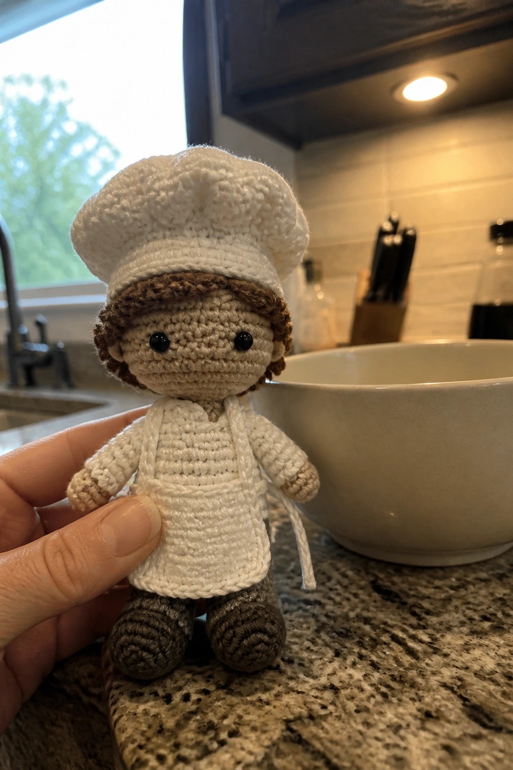 Hand holding crocheted chef doll with white hat, apron, and gray pants on counter.