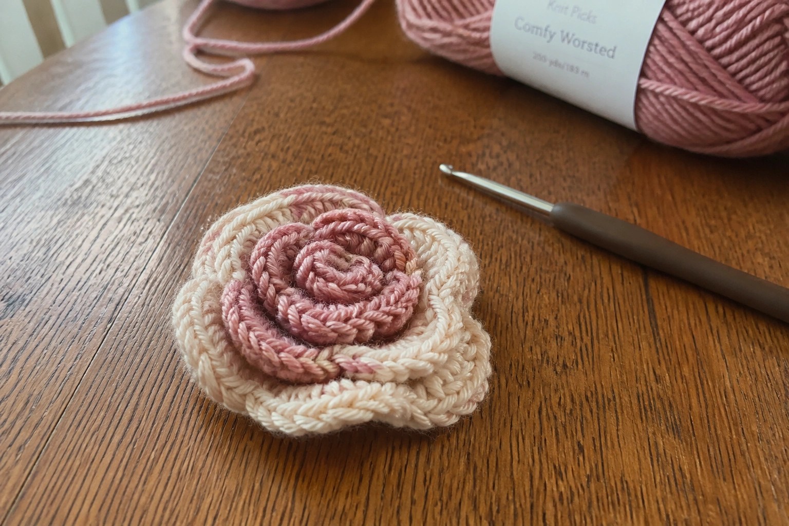 Crocheted pink and cream rose on wooden table with yarn and hook