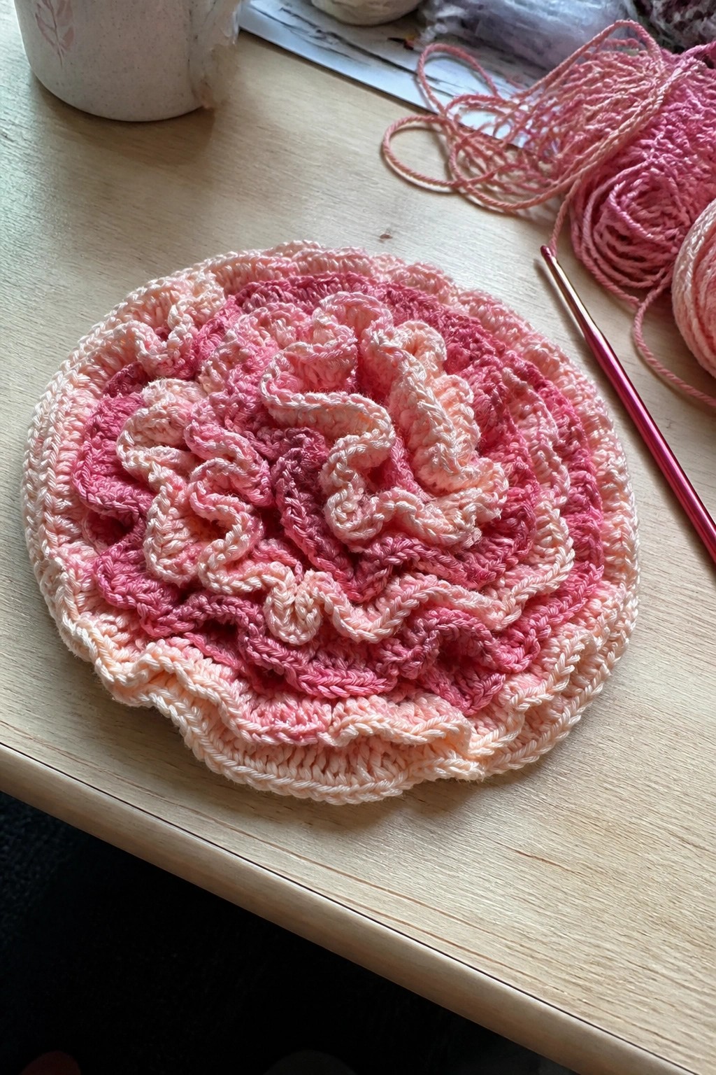 Pink gradient crocheted floral doily on wooden table with yarn and hook