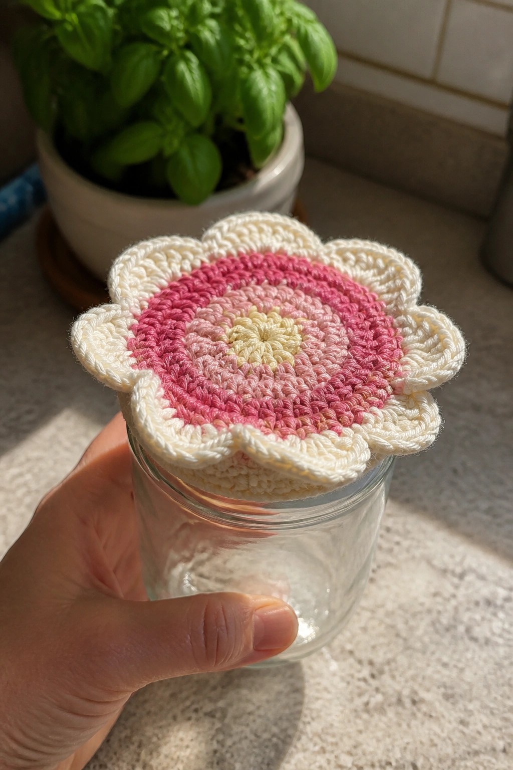 A hand holding a clear glass jar with a pink and cream crocheted flower-shaped lid cover, next to a potted basil plant.