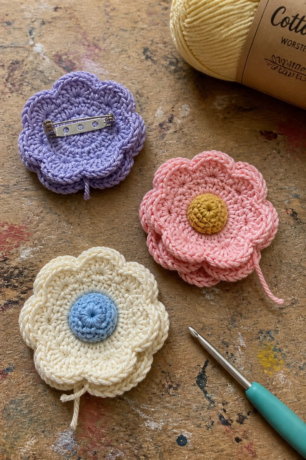 Three crocheted flower motifs in purple, pink with yellow center, and cream with blue center sit beside a crochet hook and ball of yellow yarn on a wooden surface.