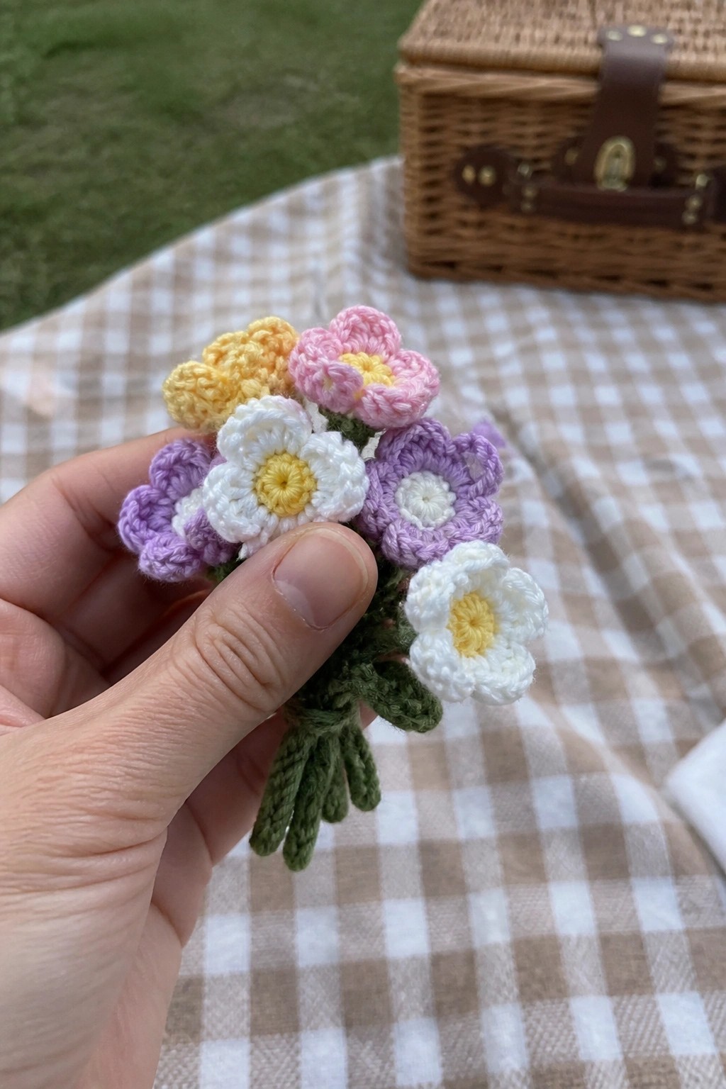 Hand holding colorful crocheted flowers over gingham picnic blanket with basket nearby